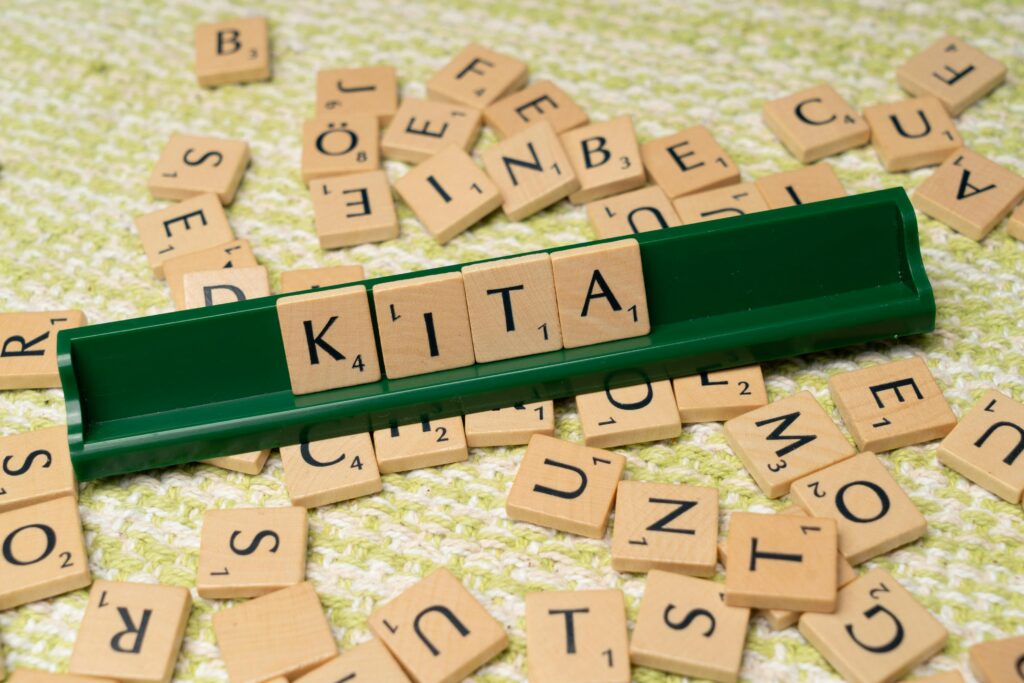 Scattered wooden Scrabble tiles with a green holder displaying the word 'KITA' on textured fabric surface.