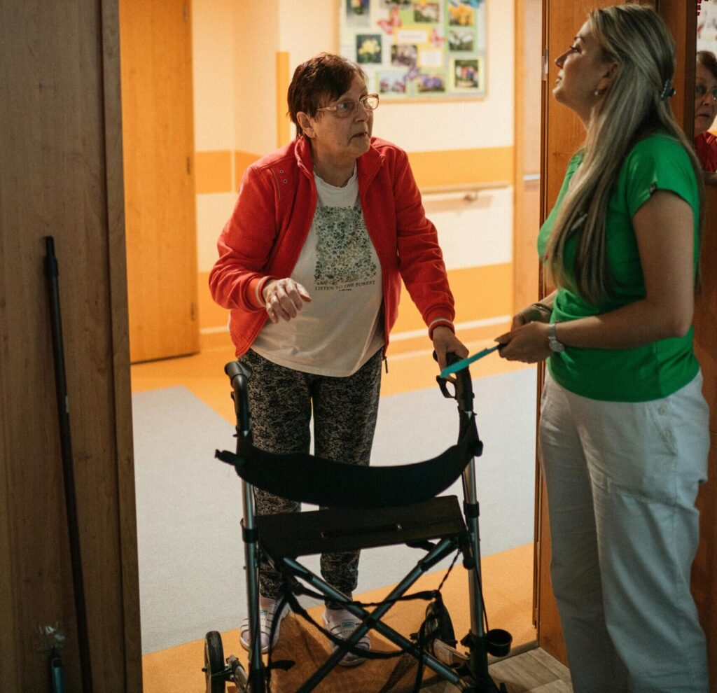 A caregiver assists a senior woman with a walker in a retirement home corridor.